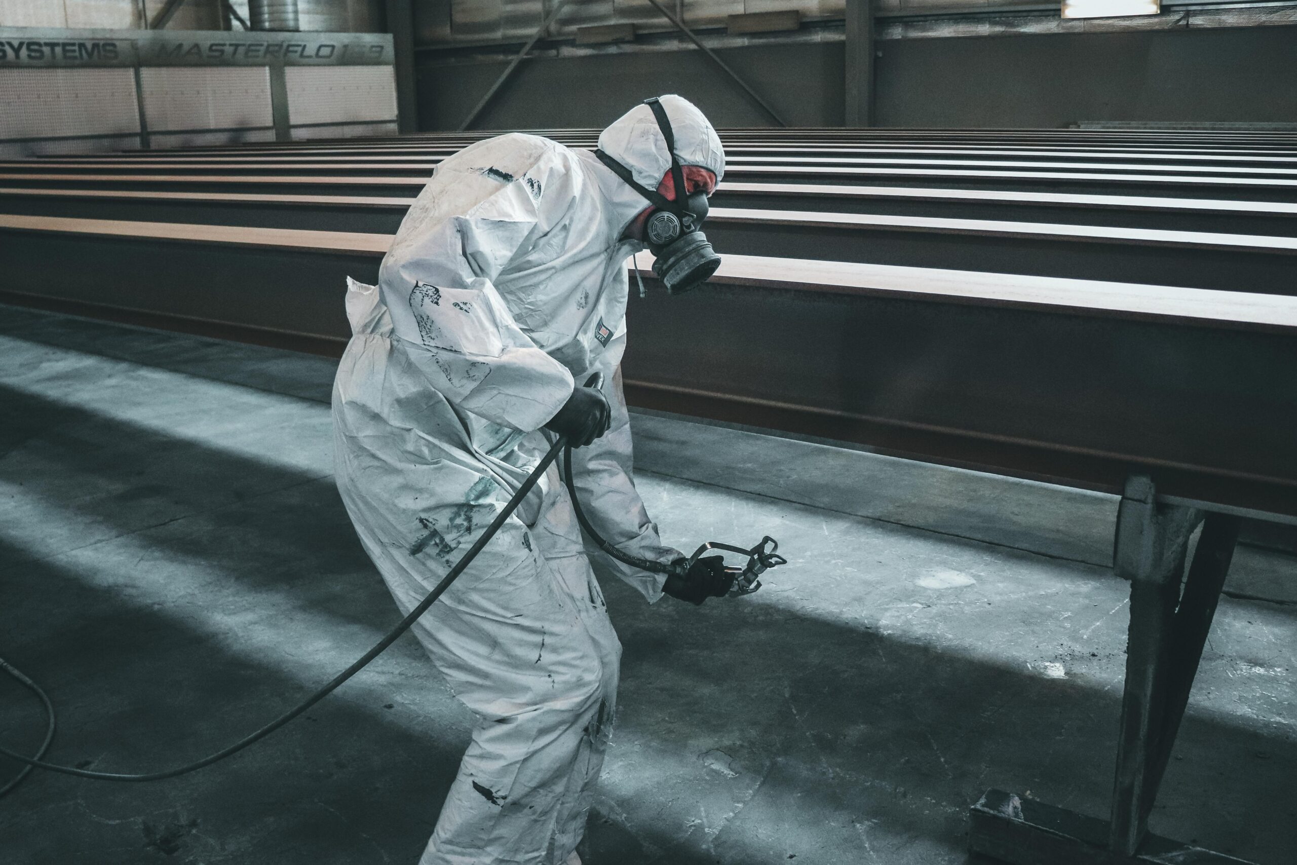 Worker in protective gear spray painting metal beams inside a factory setting.